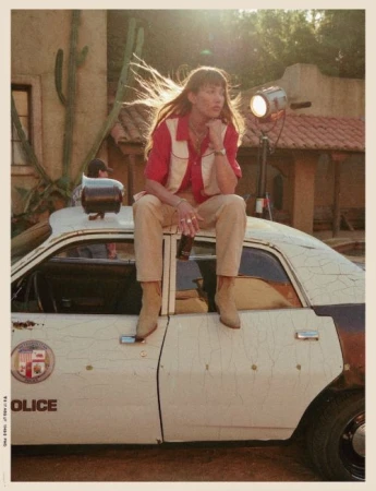 Woman with long hair sits on a weathered police car in a desert-like setting, illuminated by a light stand and backed by a building.