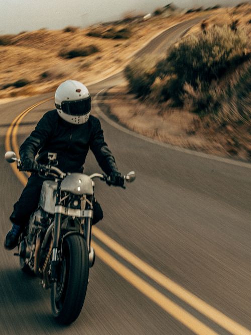 Motorcyclist riding on a winding asphalt road through a dry, hilly landscape with a helmet and leather jacket, captured in motion.