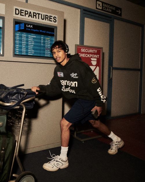 Young man with headphones pushing luggage cart in airport terminal near departure board and security checkpoint sign indoors.