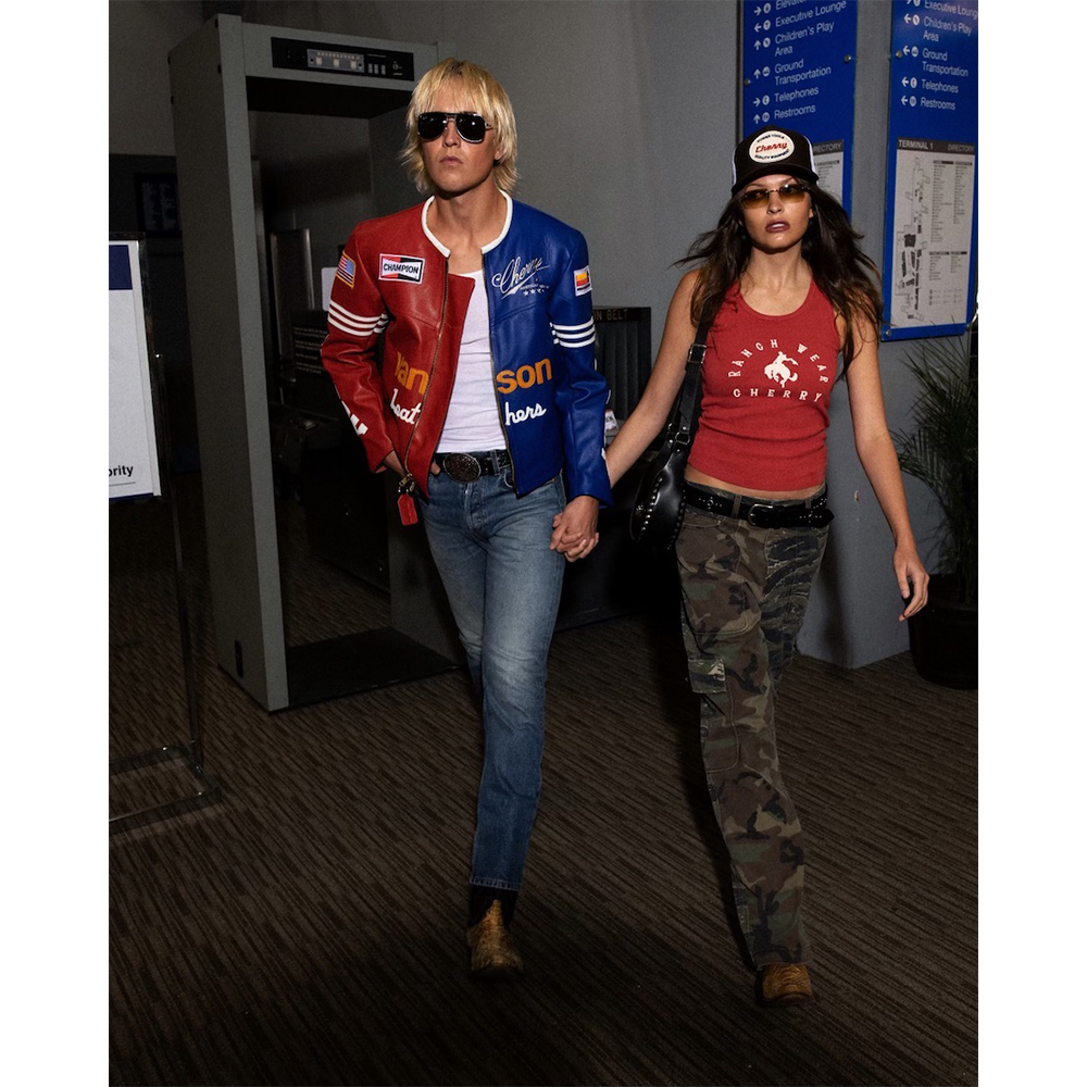 A stylish couple walking through an airport or transit hub with luggage, featuring a man in a red and blue jacket and sunglasses.