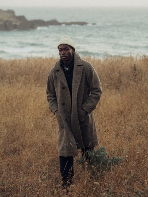 A dark-skinned man in a long wool coat and beanie stands in a field of brown grasses near a rocky coast and ocean under an overcast sky.