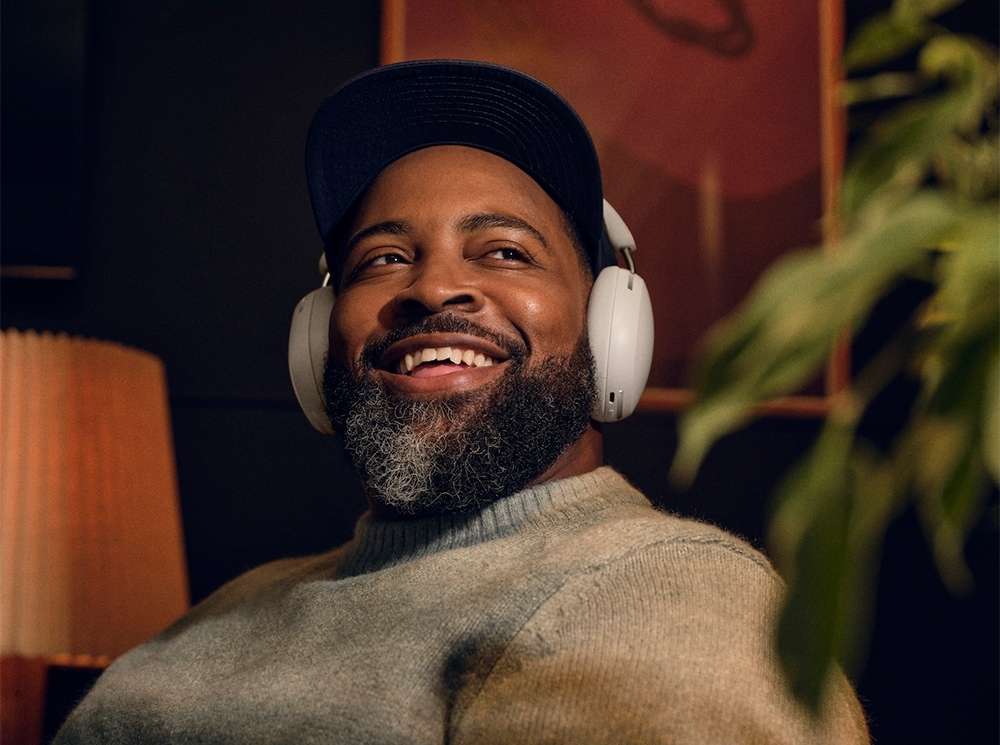 Smiling Black man with gray beard wearing wireless headphones indoors, with cap, gray sweater, and a plant in the foreground.