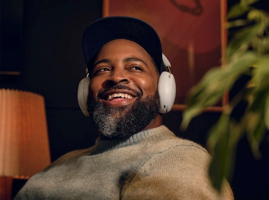 Smiling Black man with gray beard wearing wireless headphones indoors, with cap, gray sweater, and a plant in the foreground.