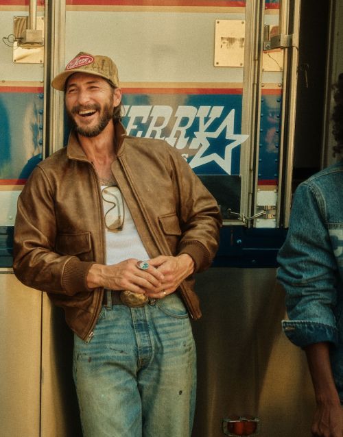 Laughing man wearing a trucker hat and leather jacket leans against a blue and white service truck with another person alongside him.
