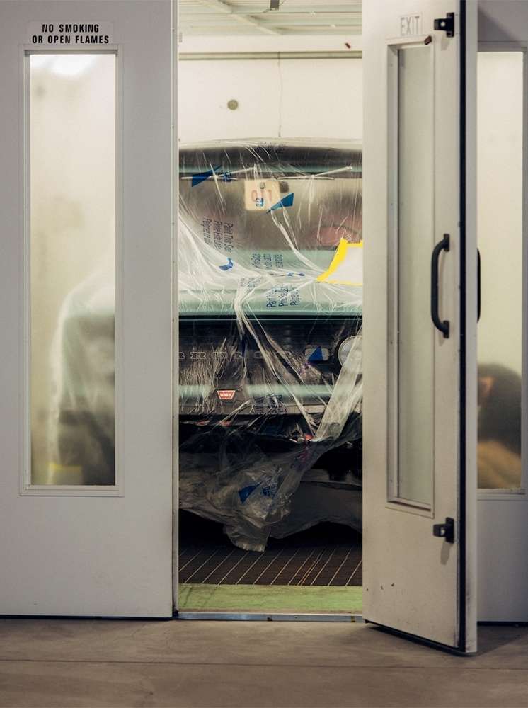 Classic car inside a paint booth, covered with plastic, doors ajar, warning sign visible. Automotive restoration project.