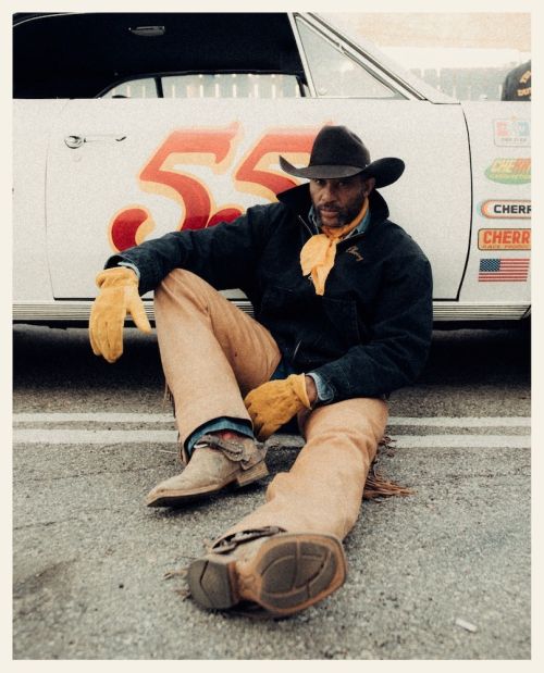 Man in cowboy attire sits by a vintage race car on an asphalt track, sporting a cowboy hat, neck scarf, and boots for an editorial look.