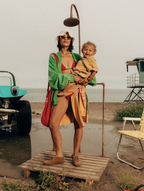 Woman in bikini holds a baby on a beach with a buggy, shower, and lifeguard tower in the background. Lifestyle fashion shot outdoors.