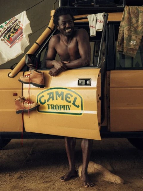 Bare-chested man smiles next to a yellow off-road vehicle with hanging work boots and clothing in an outdoor setting with a sand surface.