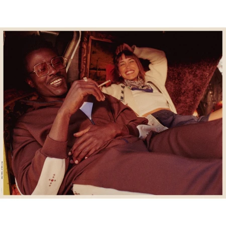 Man wearing brown tracksuit and tinted glasses sitting beside a smiling woman in a retro interior setting