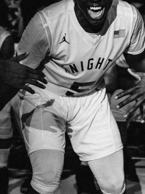 A black and white image featuring a basketball player in uniform, yelling with excitement while being supported by teammates.