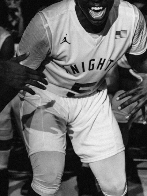 A black and white image featuring a basketball player in uniform, yelling with excitement while being supported by teammates.
