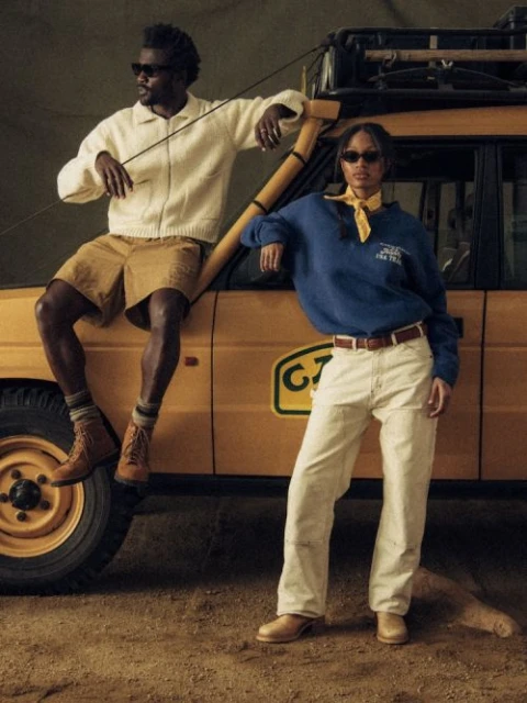 Two adult models, a man and a woman, pose next to a vintage yellow SUV outdoors with tan clothing, sunglasses, and a casual fashion style.