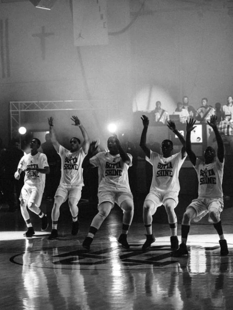Five men in matching shirts and shorts are on a basketball court with their arms raised under bright lights in a black-and-white photograph.
