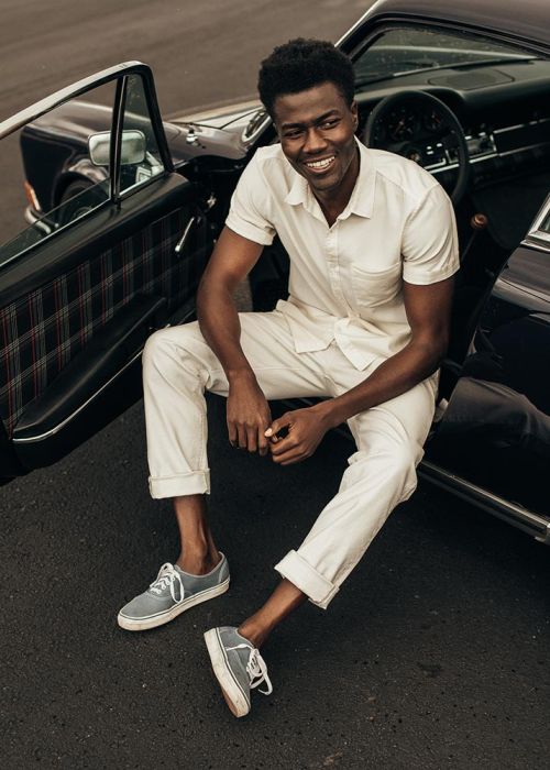Smiling African-American man in casual wear sitting on car door, with vintage car, wearing sneakers in an outdoor environment.