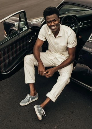 Smiling African-American man in casual wear sitting on car door, with vintage car, wearing sneakers in an outdoor environment.