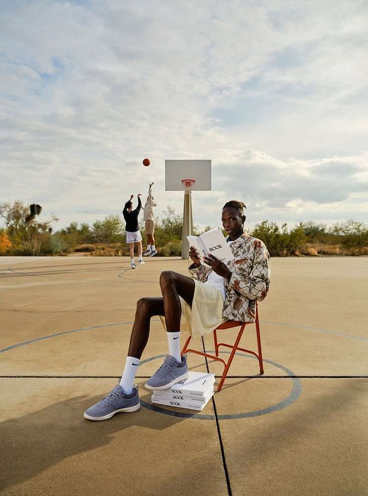 Adult male sitting on a chair, reading a book on a basketball court outdoors. Two other men are playing basketball in the background.