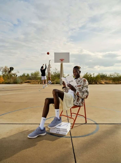 Adult male sitting on a chair, reading a book on a basketball court outdoors. Two other men are playing basketball in the background.