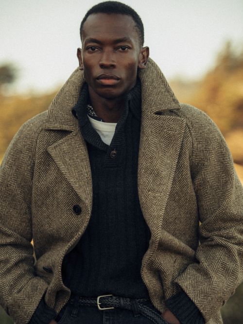 A dapper black male model poses in a tweed overcoat and dark sweater outdoors against a muted, warm-toned natural backdrop.