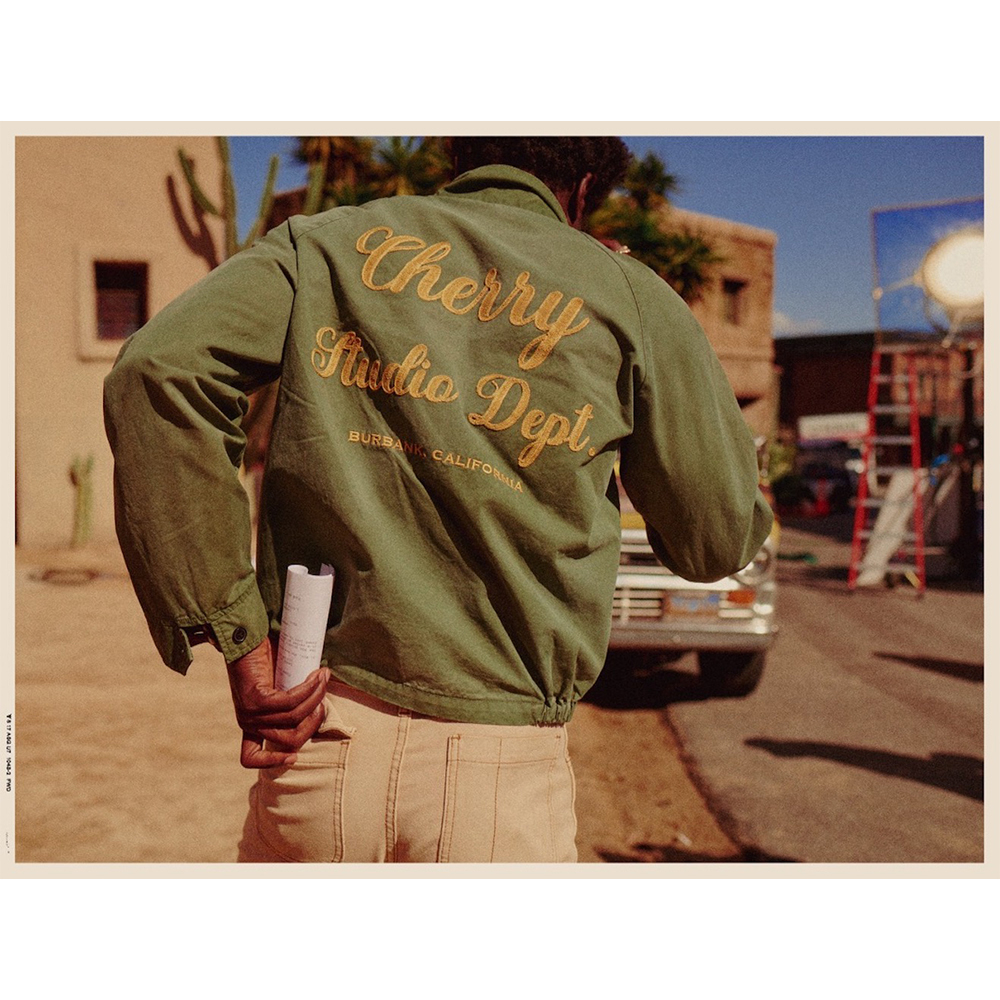 Man in olive jacket with 'Cherry Studio Dept.' text, walking on outdoor film set with vintage truck, ladders, and desert plants.