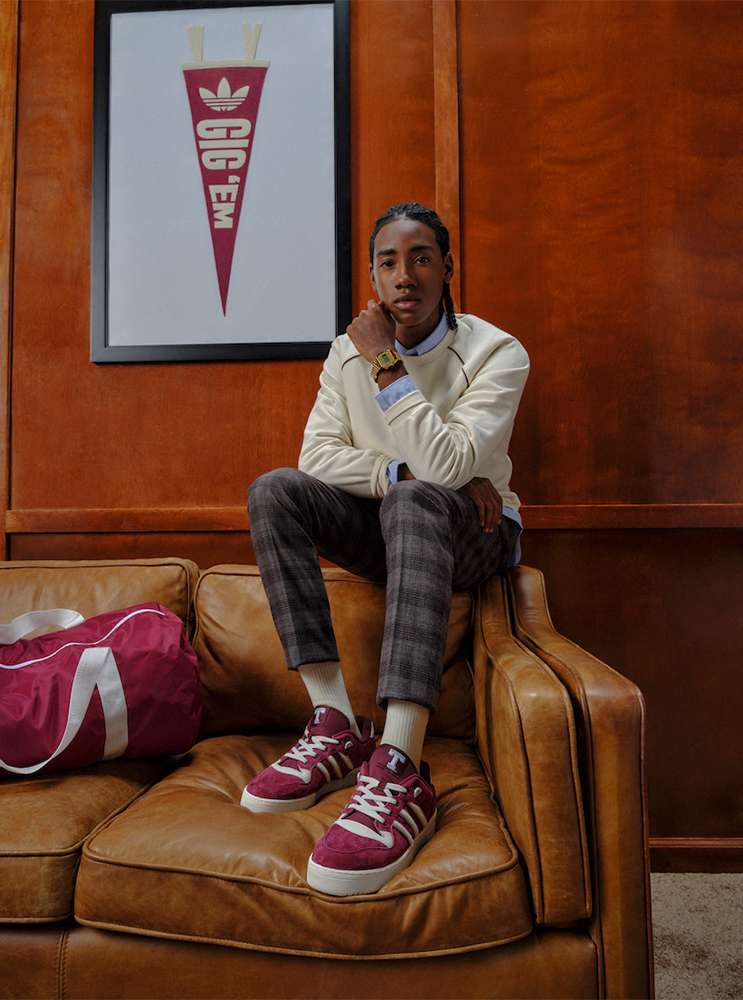 Young adult poses on a brown leather couch in casual clothing with sneakers, a burgundy duffel bag, and an Adidas pennant poster.