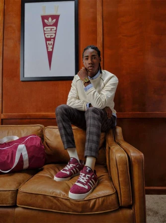 Young adult poses on a brown leather couch in casual clothing with sneakers, a burgundy duffel bag, and an Adidas pennant poster.