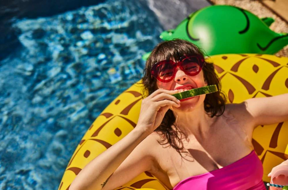 Woman in a pink tube top eating watermelon on a pineapple pool float in a swimming pool, wearing red sunglasses.