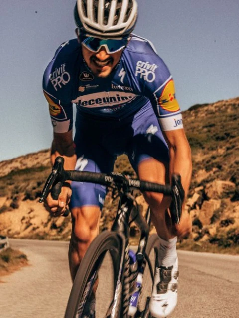 A cyclist in blue uniform races uphill on an asphalt road, helmeted and wearing sunglasses, with a clear sky and hilly landscape.
