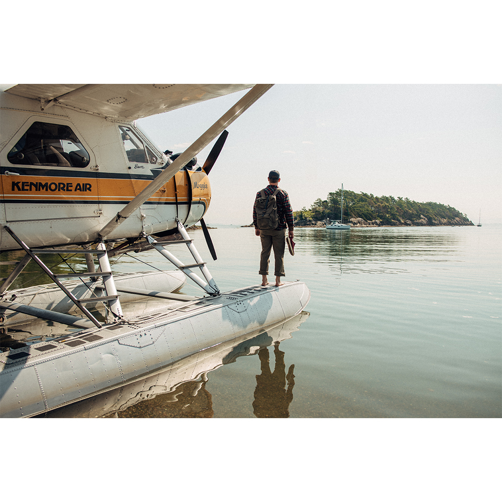 Man standing on a seaplane pontoon in water near an island. The seaplane is yellow and white, with the man facing away from the camera.