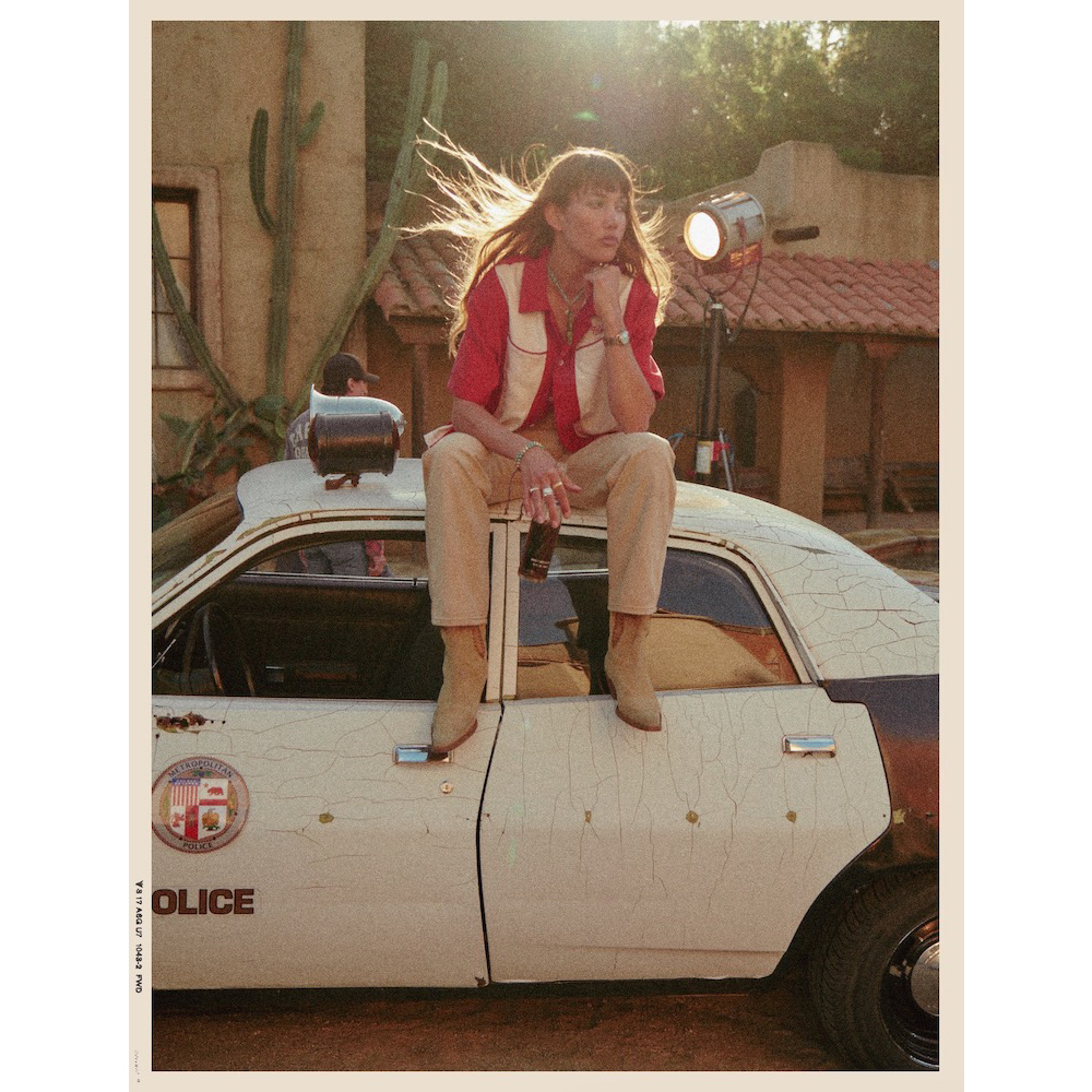 Woman with bangs sits on a retro police car outside. She wears a red shirt and cowboy boots. Desert plants and a building are behind her.
