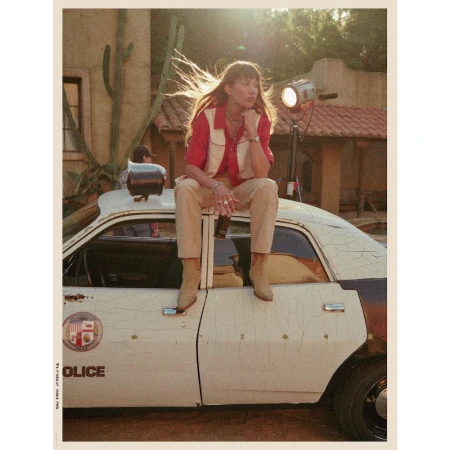 Woman with bangs sits on a retro police car outside. She wears a red shirt and cowboy boots. Desert plants and a building are behind her.