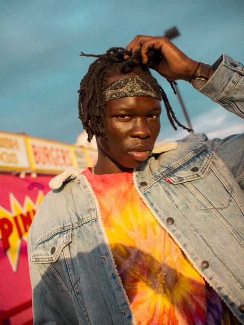 Portrait of a young man with dreadlocks and a patterned bandana, wearing a denim jacket and tie-dye shirt, standing outdoors.