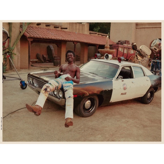 Shirtless man poses on vintage police car with desert backdrop. He wears printed pants and cowboy boots with film studio lighting.