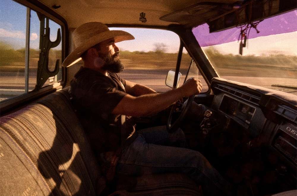 Bearded man with cowboy hat driving old pickup truck on highway during sunset, creating a vintage automotive lifestyle moment.