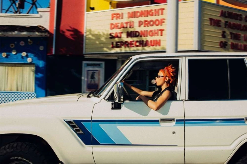 Adult woman driving a white car with blue trim while blowing a bubble gum bubble; outdoor scene, with a theater in the background.