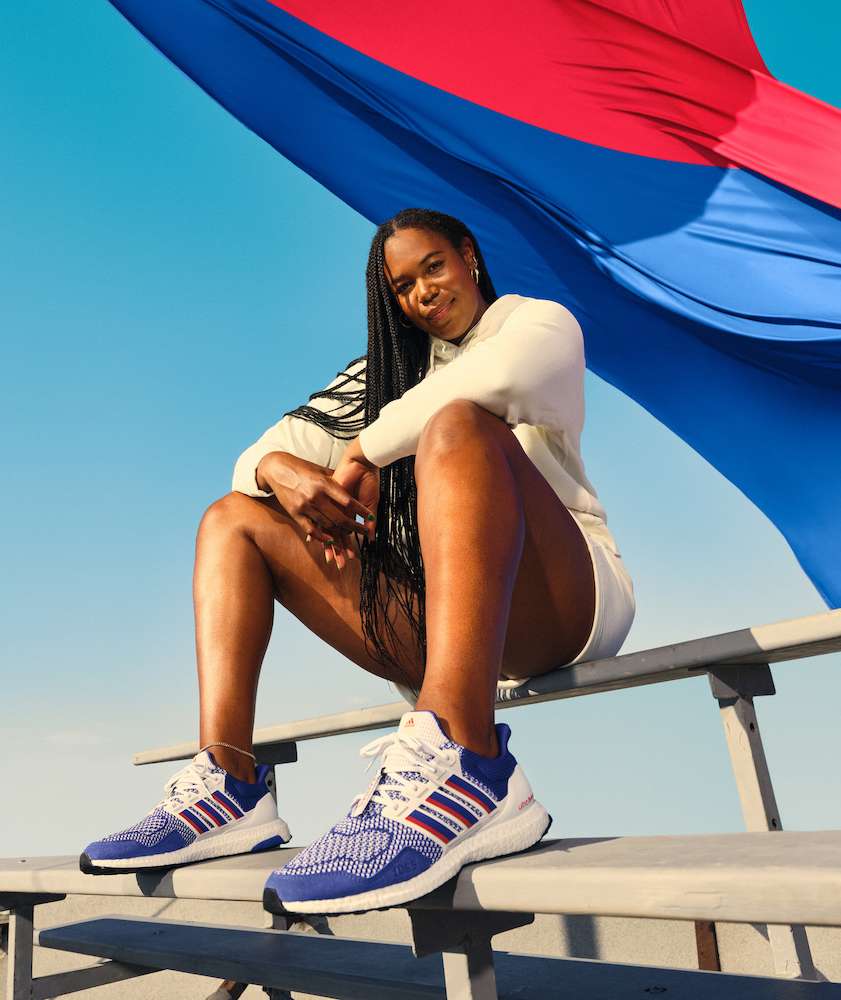 A Black woman wearing athletic shoes sits on bleachers with a blue and red flag behind her, posing in a medium outdoor shot.