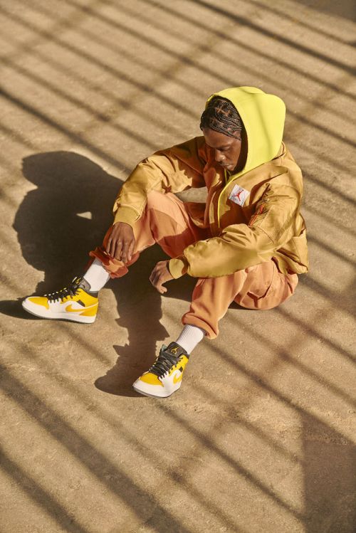 Man sits on the ground in a yellow hoodie and matching shoes, sunlight stripes across him and the pavement, casual, outdoor.