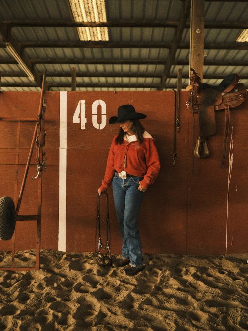 Woman in cowboy hat stands in indoor riding arena wearing jeans and red jacket holding horse lead with saddle and equipment in background.