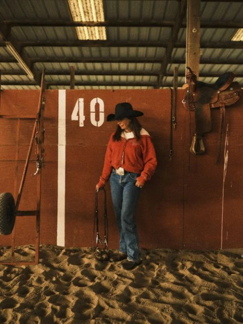 Woman in cowboy hat stands in indoor riding arena wearing jeans and red jacket holding horse lead with saddle and equipment in background.