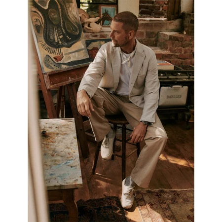 Adult man wearing a blazer and khaki pants sitting on a chair in an artist's studio with paintings and a vintage oven.