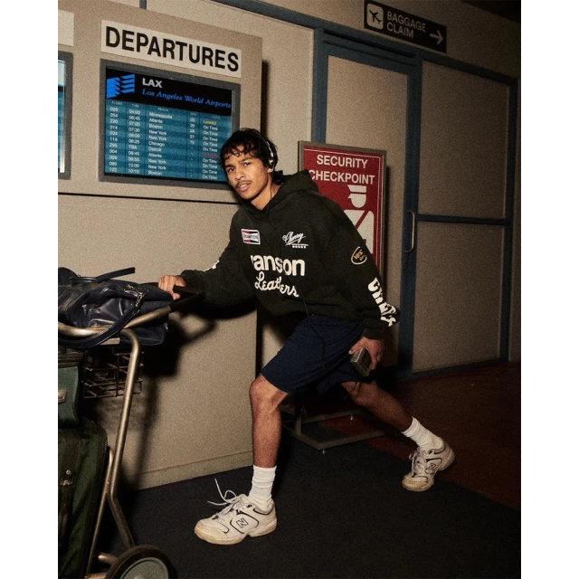 Man in airport with baggage cart and headphones walking past Departures monitor and Security Checkpoint sign in warm, diffused lighting.