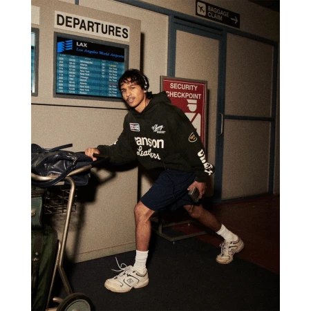 Man in airport with baggage cart and headphones walking past Departures monitor and Security Checkpoint sign in warm, diffused lighting.