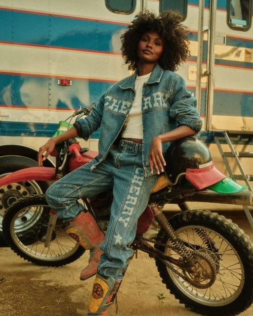 An adult female wearing denim apparel poses on a motorbike in front of a vintage-style trailer in an outdoor setting with warm lighting.