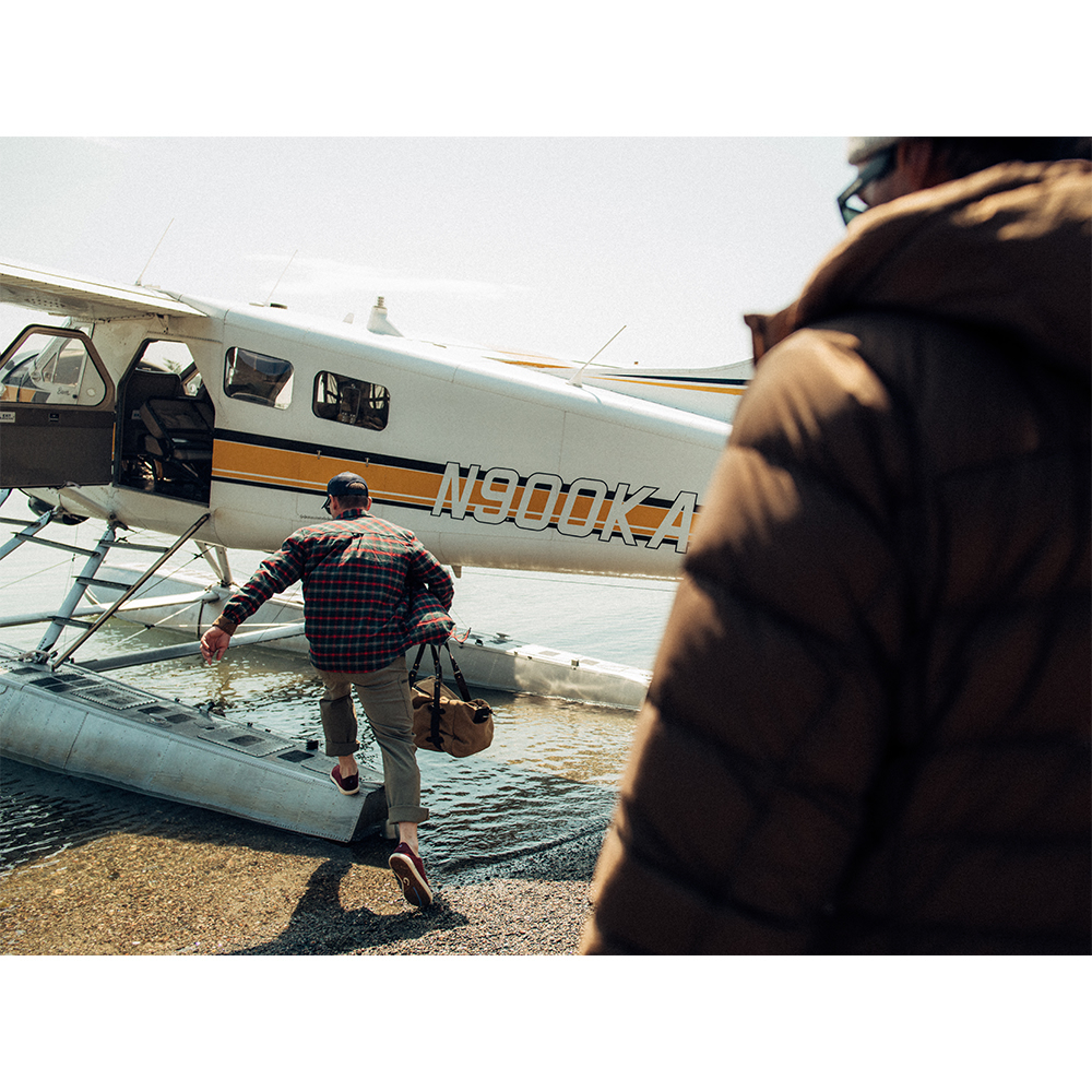 Man in a red plaid shirt exits a small floatplane, carrying a duffel bag, stepping into shallow water on a sunny day.