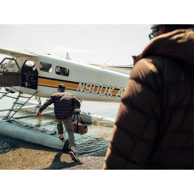 Man in a red plaid shirt exits a small floatplane, carrying a duffel bag, stepping into shallow water on a sunny day.