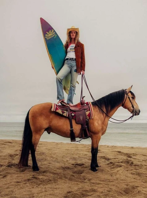 A woman in a cowboy hat stands barefoot on a horse with a surfboard at the beach on a cloudy day.