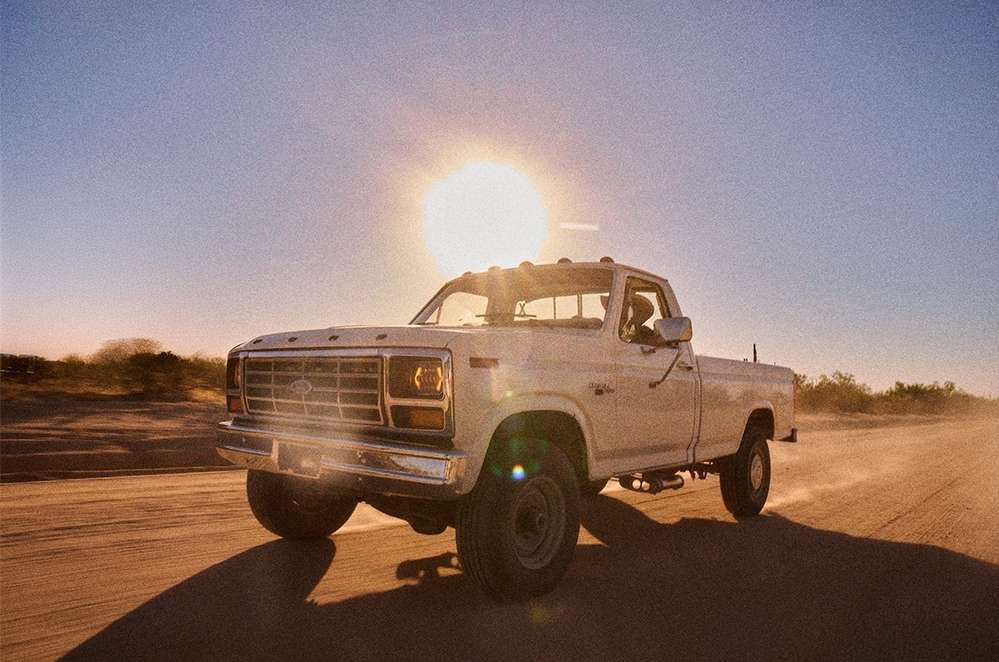 Vintage white pickup truck driving on a dusty desert road during a bright sunset by Nicholas Maggio.