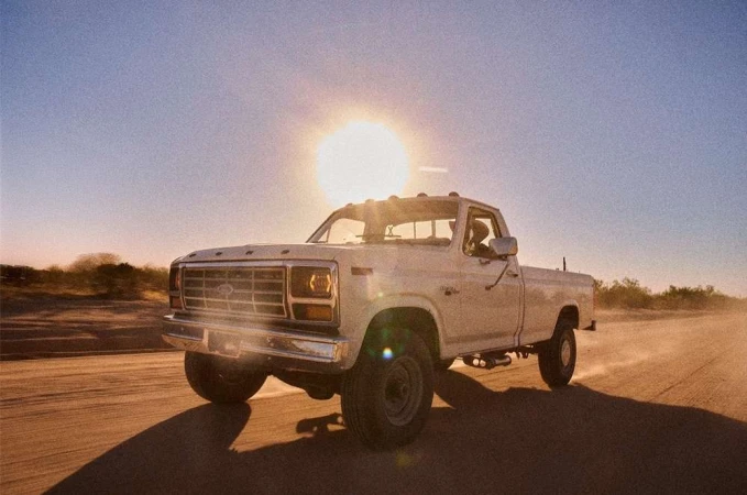 Vintage white pickup truck driving on a dusty desert road during a bright sunset by Nicholas Maggio.