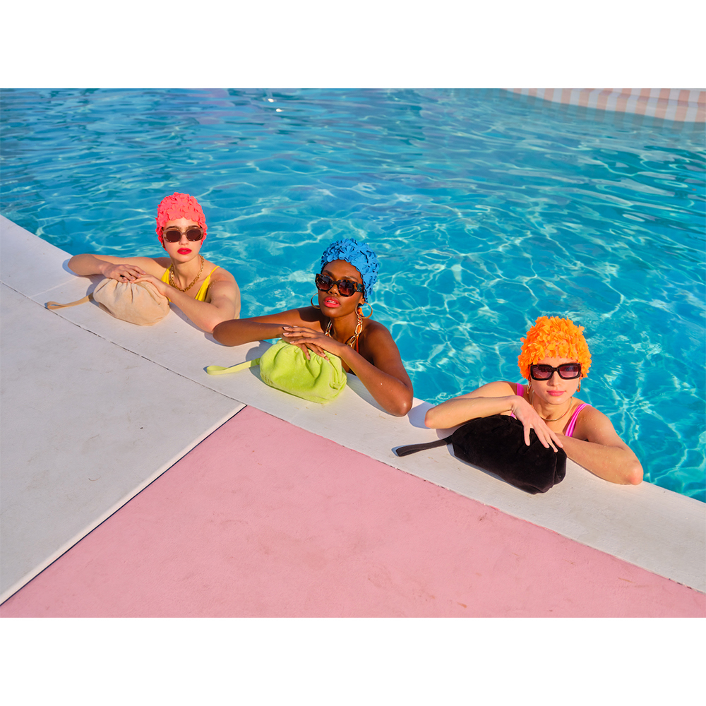 Three women in a swimming pool wear retro bathing caps and sunglasses, holding clutch bags, under bright sunlight and turquoise water.
