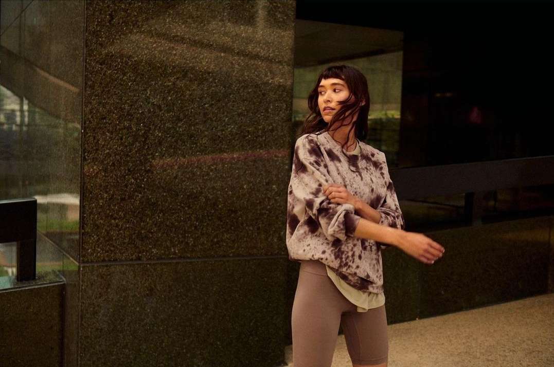 A young adult woman with dark hair and bangs poses outdoors wearing a tie-dye sweatshirt and biker shorts in front of a building.