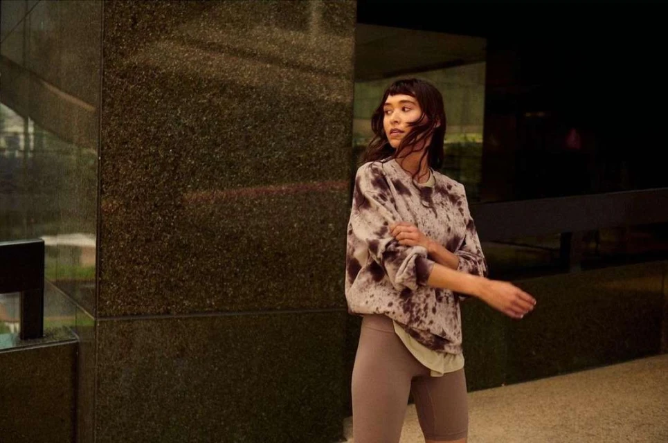 A young adult woman with dark hair and bangs poses outdoors wearing a tie-dye sweatshirt and biker shorts in front of a building.